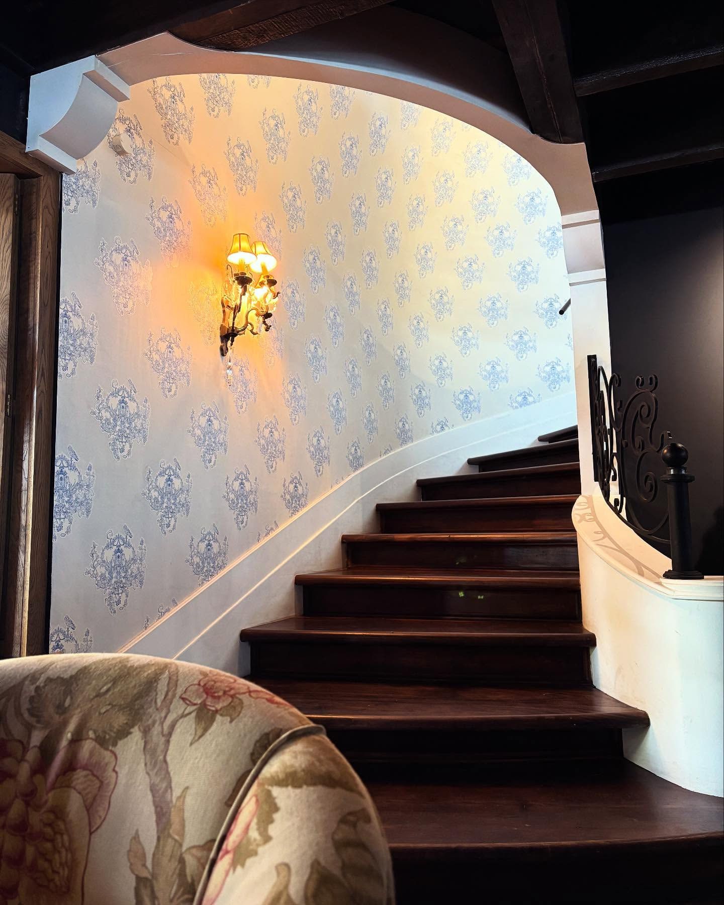 Blue and white damask wallpaper curving up a staircase lit by brass wall lamps