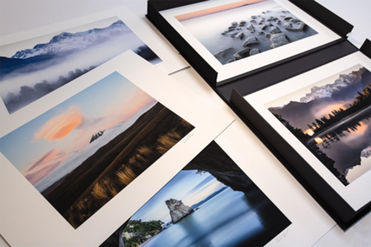 Signed fine art prints of mountains, a misty lake and a sea arch, with white borders, beside a black presentation box