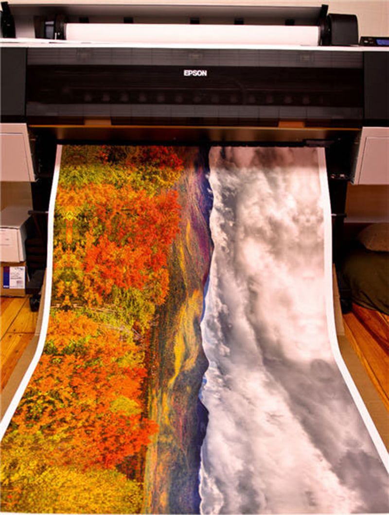 Long print of an autumn valley with orange trees and heavy grey clouds coming out of a wide format printer
